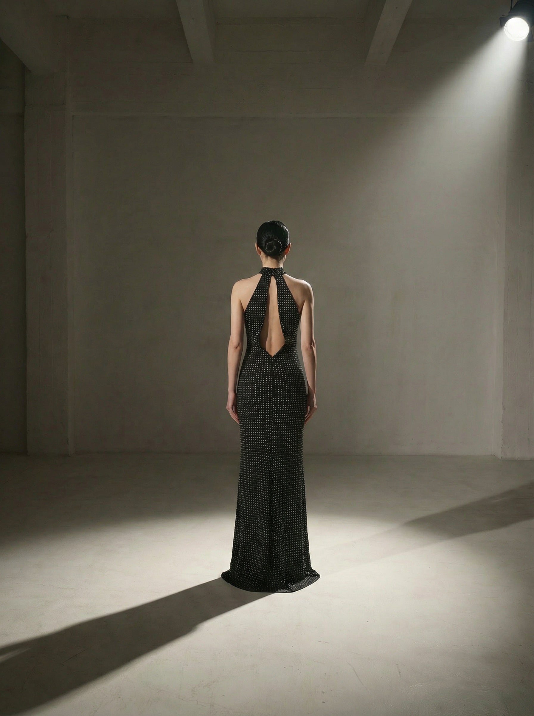 Brunette model stands centered in a shadowed room wearing a silk‑lined bias‑cut halter gown with a choker neckline, open back, and size‑2 fit.