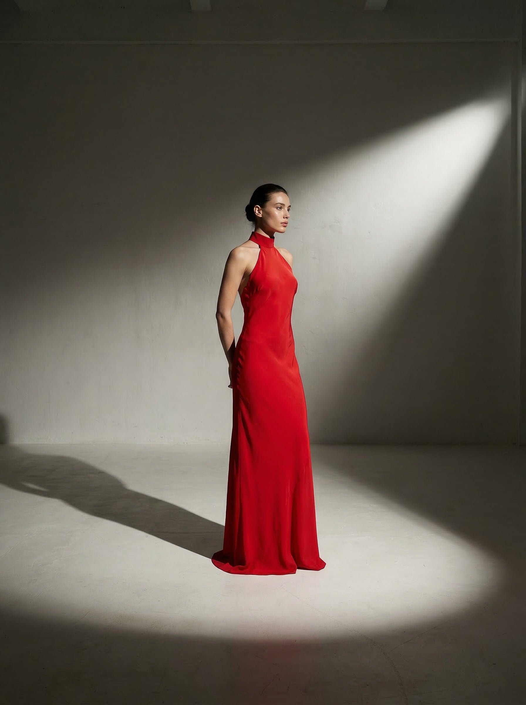 Brunette model standing in the center of a shadowed room wearing a bias‑cut halter neck gown with a close‑out choker neckline secured by two adjustable buttons and a striking open back. The gown is fully lined in silk. Model is 176 cm (5’9”) and wearing a size 2.