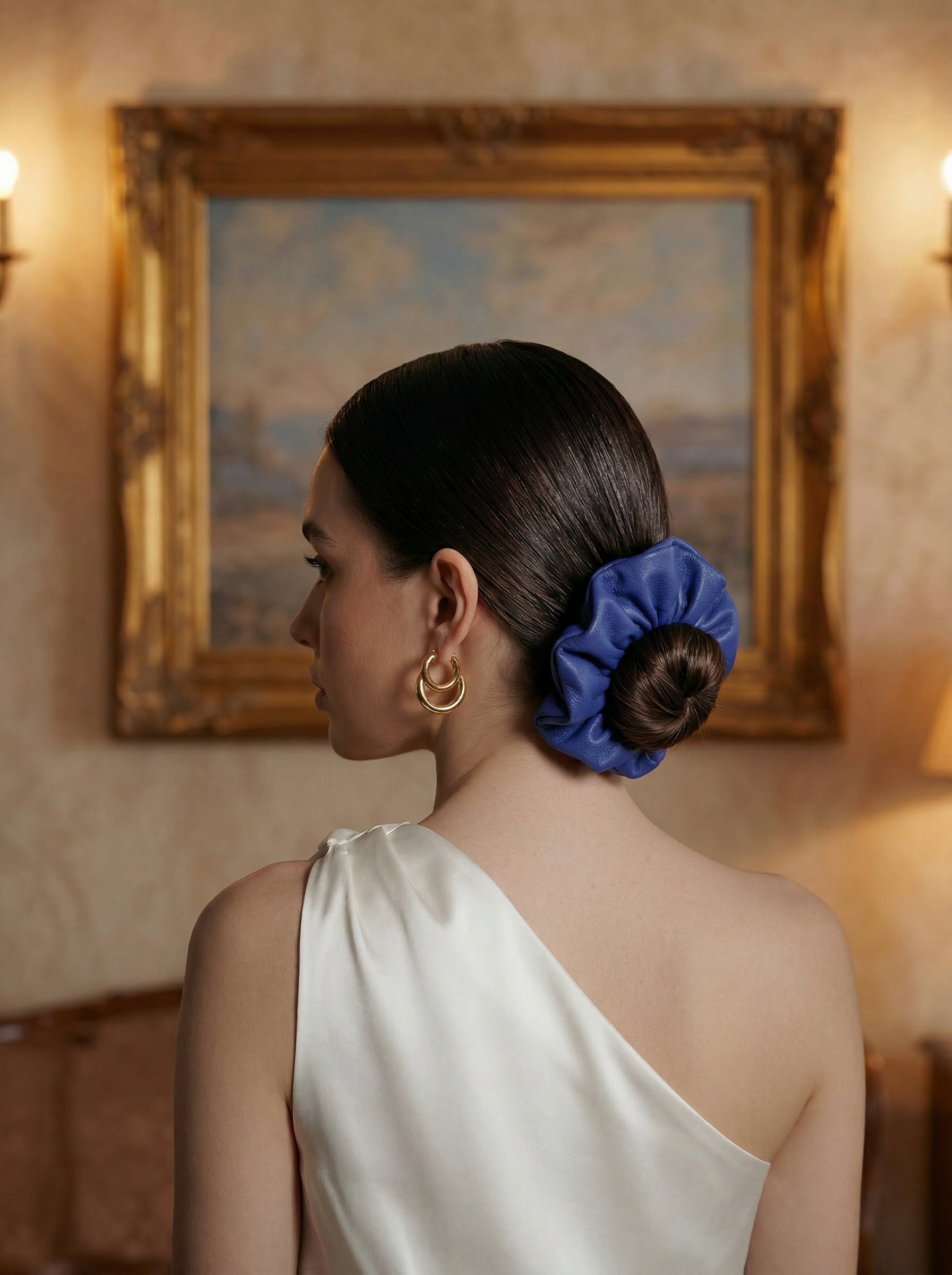 Woman with a cobalt leather scrunchie in her hair, wearing a white one-shoulder dress, in a room with a painting on the wall.