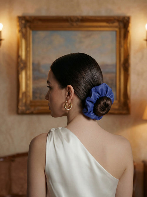 Woman with a cobalt leather scrunchie in her hair, wearing a white one-shoulder dress, in a room with a painting on the wall.