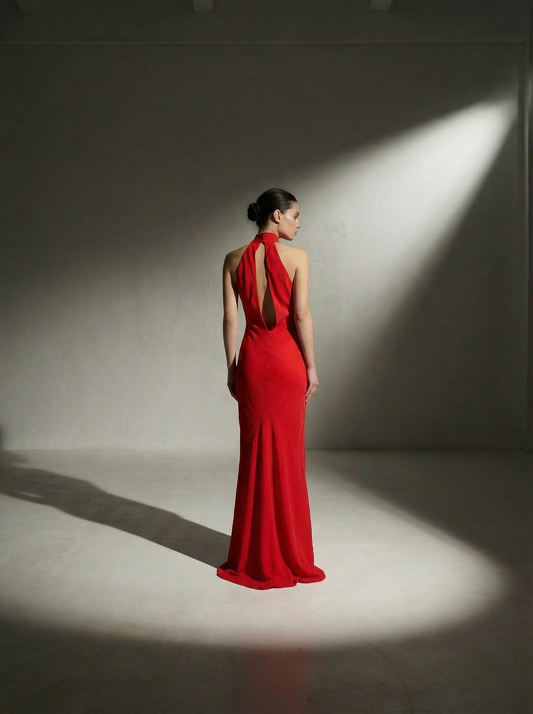 Brunette model standing in the center of a shadowed room showcasing the back of a bias‑cut halter neck gown with a close‑out choker neckline secured by two adjustable buttons and a striking open back. The gown is fully lined in silk. Model is 176 cm (5’9”) and wearing a size 2.