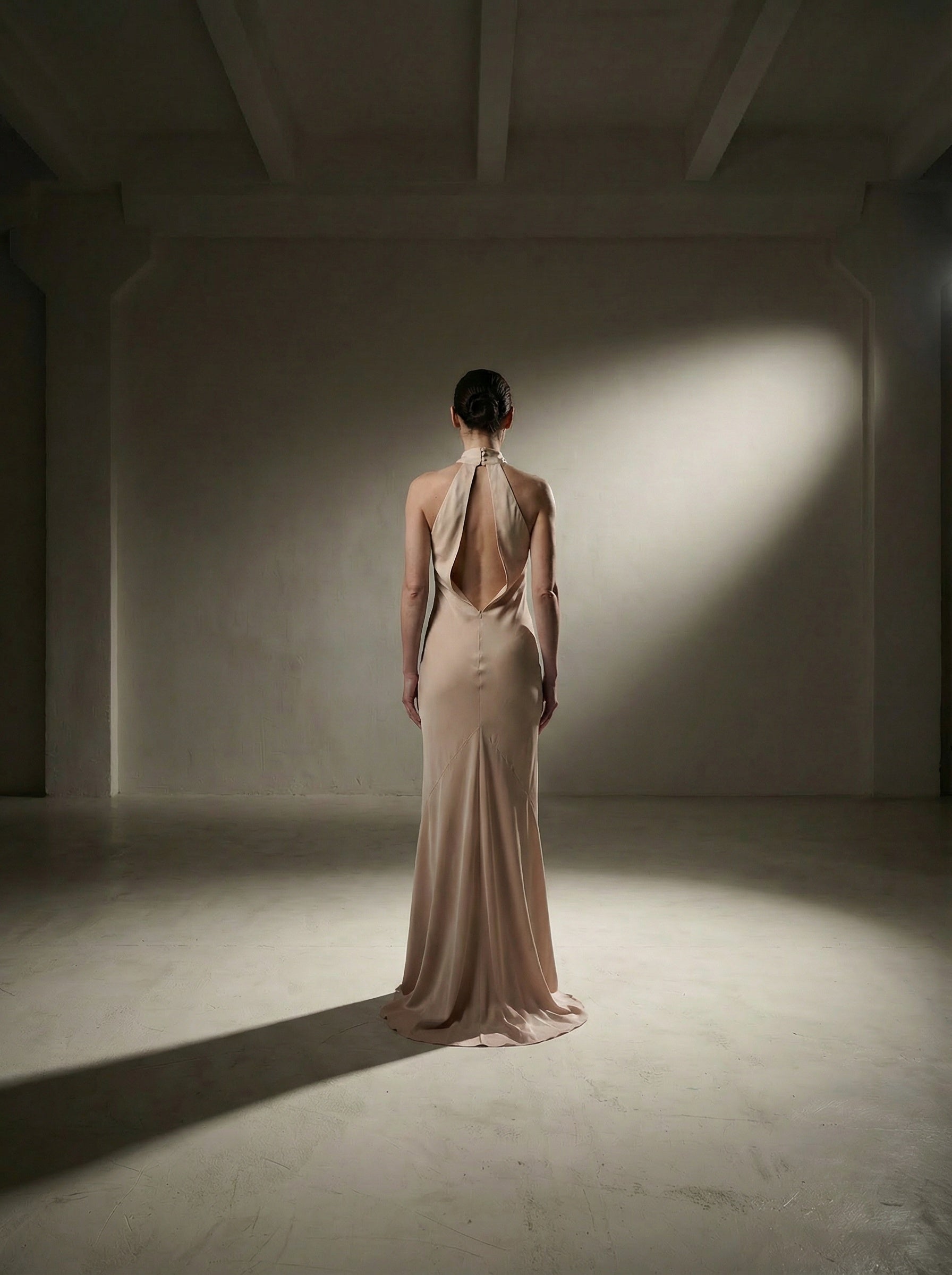 Woman in a beige evening gown standing in a dimly lit room with a spotlight on her.